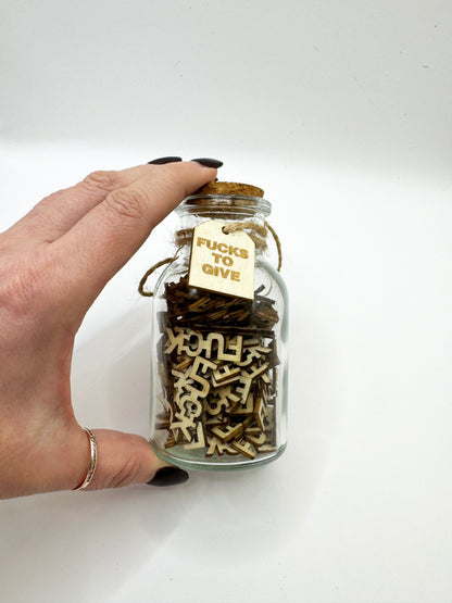 Hand holding The Swear Jar filled with wooden letters, including a 'FUCKS TO GIVE' tag.